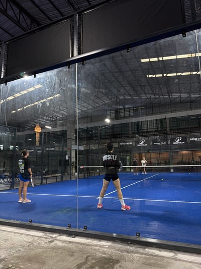 The image depicts an indoor padel tennis court in Ban Buak Khrok North, Thailand. Four individuals are visible on the blue court, engaged in a game. Two women, dressed in athletic wear, are on one side of the net, with one in the foreground facing away from the camera, holding a racket. The other two players are on the opposite side of the net, also participating in the match. The court is enclosed by glass walls and metal fencing, with a netted ceiling above. Banners bearing logos such as "Niko Padel Academy" and "Decathlon" are displayed on the back wall. The lighting suggests an indoor facility, with overhead lights illuminating the playing area. The surrounding environment appears to be a sports complex, with seating areas and other courts possibly present in the background.