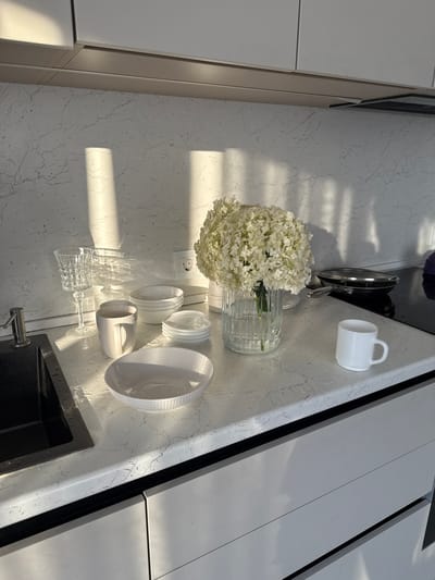 A sunlit kitchen counter in Nizhneyarkeyevo, Russia features a variety of white tableware and a vase of white flowers. On the left, a black sink is visible with a silver faucet. To the right of the sink, a clear crystal wine glass, a ribbed white mug, and several stacked white bowls are arranged. In the center, a large clear glass vase holds a bouquet of white hydrangeas. To the right of the flowers, a small white mug sits on the counter. In the background, a black stovetop with a frying pan is visible. The countertop is white with a subtle gray veining pattern, and light streaks across it from the sun.