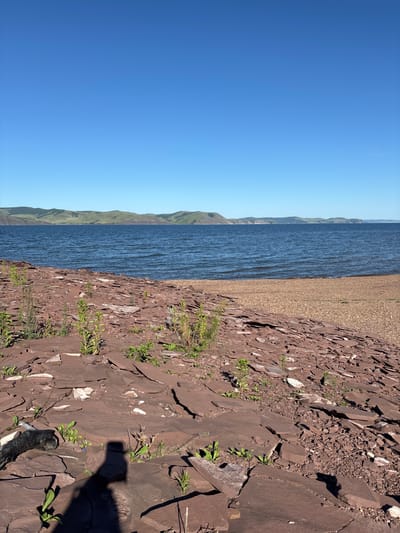 The image depicts a wide, tranquil body of water with a clear blue sky overhead. In the foreground, a rocky shoreline is covered in flat, reddish-brown stones and scattered green plants. The water is a deep blue, with gentle ripples suggesting a light breeze. Across the water, a chain of rolling green hills forms the distant horizon. The scene is serene and natural, possibly a park or natural reserve in Sovetskaya Khakasiya, Russia. A shadow of a person taking the photograph is visible in the lower right portion of the frame.