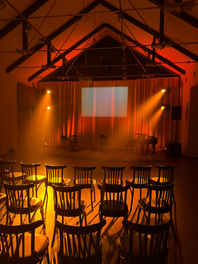The image depicts an empty auditorium or performance space, bathed in warm, orange-toned lighting. Rows of dark wooden chairs with dark upholstery face a stage area. The stage is set with a grand piano, a music stand, and a screen. The floor in front of the stage is decorated with numerous small, white objects, possibly candles or floral arrangements, creating a circular border. The ceiling features exposed beams and industrial lighting fixtures. The overall atmosphere is atmospheric and theatrical, suggesting an event is about to begin or has recently concluded. There are no people visible in the scene. The location in the city of Ilyanskiy selskiy Sovet, Belarus is not ascertainable from the visual information provided in the image.