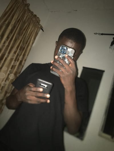 A man in a dark t-shirt holds two phones. One phone is held up to his face as if taking a picture and has a camouflage case. The other is held in his other hand and has a textured black case with what appears to be a white sticker on the back. He is in a room with golden curtains to the side and an air conditioning unit in the background. The image is rotated, and the lighting is dim.