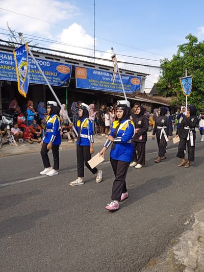 The image captures a parade procession in Winong, Indonesia, on what appears to be a sunny day. The focal point is a group of young women marching on the asphalt road. The women at the front of the procession wear matching blue jackets with yellow accents, black pants, white hats, and carry flags on poles. They wear sneakers. Following behind them are women dressed in black outfits cinched with white belts. These women carry pieces of cardboard in their hands and some wear sandals. Spectators, mainly women and children, are sitting and standing along the side of the road, observing the procession. In the background, storefronts bear signage in Indonesian advertising grocery and plastic goods. The signs also include phone numbers and indicate "Desa Pekalongan," which refers to Pekalongan village, near Winong. An overhead electrical line spans the street. The image suggests a local community event or celebration.