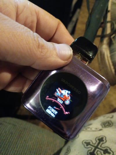A person's left hand holds a purple Vaporesso electronic cigarette. The device's circular screen displays an image of a motorcycle with the word "Vaporesso" above it, and "1660" is displayed on a gauge-like display, next to "POWER 100%." In the background, there is an armrest and a cushion with a cross pattern. The location is presumed to be Clarksville, United States, as per the instructions.