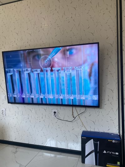 The image shows a television displaying a science experiment with test tubes and a pipette, mounted on a wall with marble-patterned wallpaper. Below the television, on the floor, is a PlayStation 5 Pro box. Two electrical outlets are visible on the wall. The floor is tiled with a marble pattern. The scene appears to be indoors, likely in a residential setting, located in Benkovski, Bulgaria. There are no people visible in the image.
