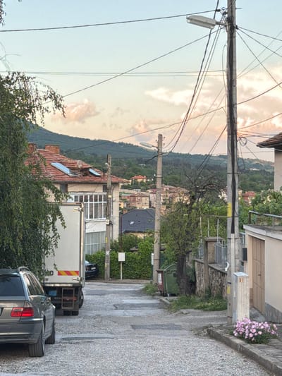 This image captures a residential street scene in Karlovo, Bulgaria, during what appears to be late afternoon or early evening. The sky transitions from a pale blue overhead to a faint pinkish-orange at the horizon, with sparse, wispy clouds. A prominent range of dark green, forested hills and mountains forms the distant background.
The street in the foreground is paved with uneven, light-colored stones or gravel and slopes upward into the distance. On the left, a dark grey station wagon (identifiable as a BMW E46 Touring by its rear design) is parked, facing away from the viewer. Immediately behind it, a white truck, likely a delivery vehicle (with "DAILY" partially visible on its rear and a yellow and red reflective strip), is also parked. Overhanging green tree branches partially obscure the upper left side of the scene.
Further up on the left, a multi-story building features a red tiled roof, a section of which appears to be damaged or partially collapsed. Two modern, rectangular skylight windows are integrated into the roof. Below the roofline, an enclosed balcony or sunroom with multiple glass panes is visible.
On the right side of the street, a tall concrete utility pole is heavily laden with numerous black electrical and communication cables wrapped vertically and extending outwards in various directions. A grey, rectangular street light fixture is mounted on this pole. Further up the street, another similar utility pole is visible. Along the right curb, a small patch of pink flowers is blooming. Two green waste bins, one with a red side, are located further along the street near a wall and a tree with bare branches. A light-colored building with a brown garage door is also present on the right.
In the mid-distance, numerous other residential buildings with red or dark tiled roofs are scattered among green trees, extending up the lower slopes of the hills. A faint, light-colored horizontal structure, possibly a road or another utility line, is barely discernible on a distant forested hill towards the upper right. No individuals or animals are visible in the scene.