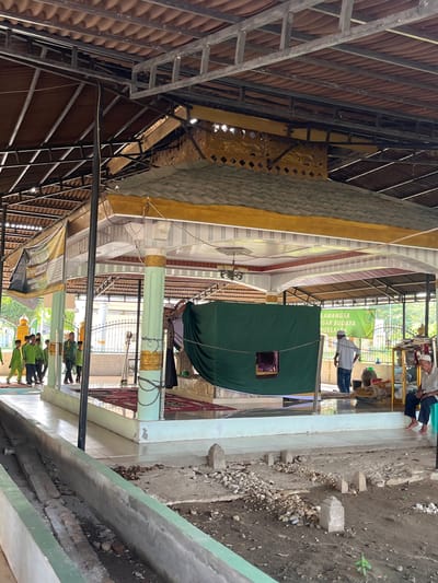 The image depicts the courtyard of a memorial or shrine, likely in Beuringen, Indonesia. The central structure is a covered mausoleum, with a green tarp draped around it. Several people are present, including a group of young males in green shirts walking in the foreground, and an older man seated to the right. The environment includes a tiled walkway, grave markers, and a metal-framed roof structure. There are also banners with text, one of which appears to be religious in nature. The scene suggests a place of religious or historical significance, possibly a pilgrimage site.