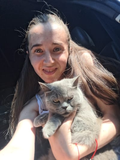 A woman with long brown hair is taking a selfie in a car. She is holding a grey cat in her arms. The cat is looking at the camera with a grumpy expression. The woman is smiling. The background is dark and blurry, suggesting the car interior. The scene is lit by sunlight, casting shadows. This photograph was taken in Vypasne, Ukraine.
