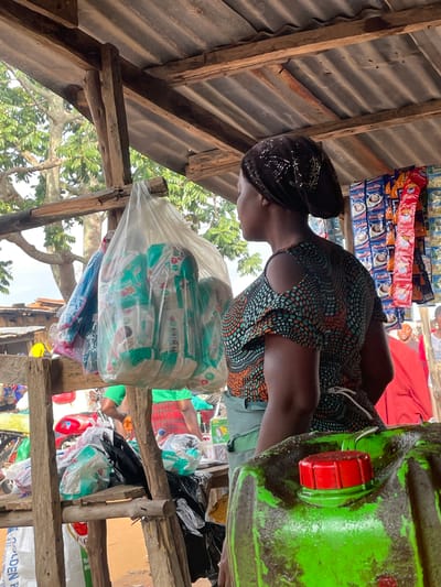 A woman, wearing a patterned dress and a beaded headscarf, stands in what appears to be an outdoor market stall in Wang, Nigeria. She is looking to her left, with her body angled slightly away from the viewer. In front of her, a large, bright green plastic container with a red cap is prominently displayed. Behind her, shelves are stocked with various packaged goods, including what seem to be sachets of coffee or other beverages, and a large clear plastic bag filled with smaller packaged items, possibly diapers or hygiene products. The stall is constructed from corrugated metal for the roof and rough wooden beams. In the background, trees and foliage are visible, suggesting a rural or semi-rural setting. The lighting suggests it is daytime, likely sunny. The overall scene depicts a vendor at her market stall, with goods ready for sale. There is no visible text in the image that is clear enough to read.