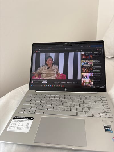 The image shows a laptop open and displaying a YouTube video. The laptop is a silver HP Pavilion Plus model. On the screen, a YouTube video is playing, featuring a young man with dark hair wearing a beige hooded sweatshirt with black stripes on the sleeves. He appears to be speaking directly to the camera, with a checkered black and white background behind him. The video title, visible on the screen, is "Papá CynFiltro - Mateo Garrido Lecca" and it's presented by "La Maquilladora & Co.". Other video thumbnails and search bars are also visible, indicating a typical YouTube interface.
To the right of the laptop screen, the keyboard and touchpad are visible. The keyboard has a light gray finish with Spanish key legends. A sticker on the bottom right of the laptop base reads "HP Pavilion Plus Laptop" and highlights features like "Mejor experiencia visual en donde quieras" (Best visual experience wherever you want), a description of the screen, camera, and sound, and mentions sustainable materials.
The scene is set in an indoor environment, likely a home, with a white, textured surface dominating the background, suggesting a bed or sofa. The lighting appears to be ambient room lighting, with no strong indications of natural light or a specific time of day. The overall mood is casual and relaxed, with the focus on the content being viewed on the laptop.