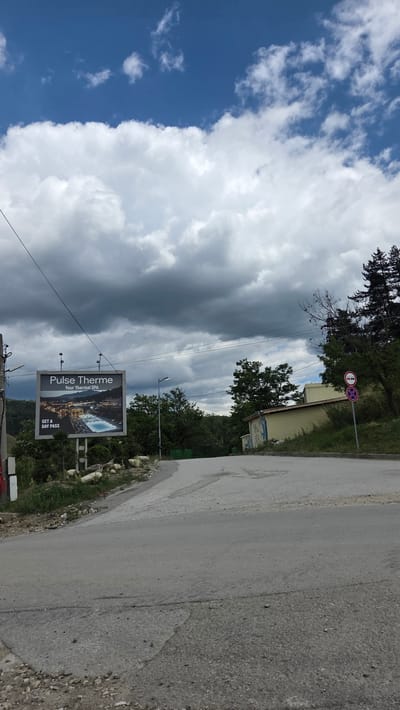 The image shows a cloudy sky over a road in the city of Bania, Bulgaria. On the left side of the road, there is a billboard advertising "Pulse Therme Your Thermal SPA" with a picture of a swimming pool. Further down the road, there are trees and a building. On the right side of the road, there is a traffic sign. The road is paved and appears to be in moderate condition, with some cracks and patches. The surrounding environment is hilly and covered with vegetation.
