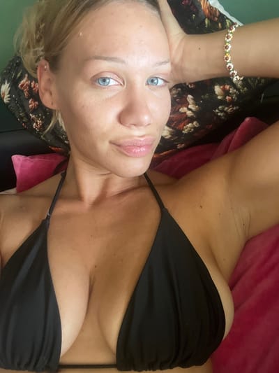 The image shows a close-up, rotated view of a person lying down. The subject has light skin, blonde hair, and striking blue eyes. They are wearing a black bikini top and a beaded bracelet with small daisy-like charms on their wrist. The background includes a dark floral fabric, a pink cushion, and a green surface, suggesting an indoor setting.

The person's face is partially visible, with their gaze directed towards the camera. Their expression appears relaxed or serene. The lighting suggests it could be daytime, possibly indoors with natural light. There is no visible text in the image. The context of Mühldorf a. Inn, Germany, provides a geographical reference but does not offer specific visual cues within the image itself. The activity appears to be resting or posing for a photograph.