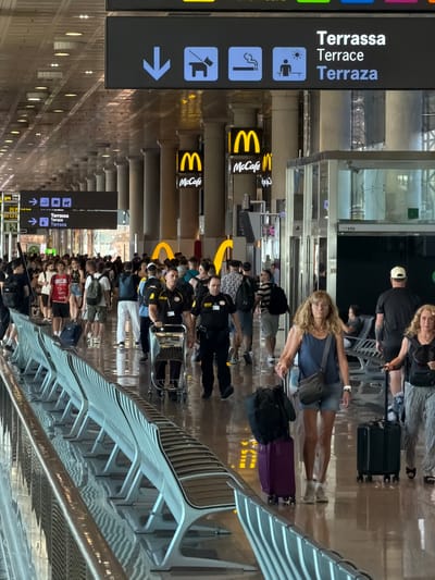 The image shows an interior view of a busy airport terminal in El Prat de Llobregat, Spain, likely Barcelona-El Prat Airport. A wide corridor with a highly reflective floor stretches into the distance, lined with rows of modern, white and gray seating. Overhead, illuminated signs indicate directions and amenities. One prominent sign reads "Terrassa Terrace Terraza" with directional arrows. Another sign displays icons for pets, smoking, and relaxation in the sun. Yellow McDonald's and McCafé logos are visible in the mid-ground, suggesting the presence of these establishments within the terminal.
Numerous people are walking through the terminal, many with luggage. In the foreground, two individuals, one female with blonde hair wearing a blue sleeveless top and denim shorts, and another female partially visible to her right, are walking with rolling suitcases. Behind them, a male airport employee in a dark uniform and a hat is pushing a luggage cart. Further back, a diverse crowd navigates the space, with individuals carrying backpacks, rolling suitcases, and some stopping to look at signs. The overall atmosphere is one of transit and travel, with the airport environment characterized by its clean, modern design and bright lighting.
