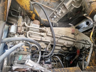 This is a close-up, high-angle, eye-level shot of the engine compartment of a heavy-duty vehicle, likely a dump truck or mining equipment, in Zheleznogorsk-Ilimsky, Russia. The engine, a beige Cummins, is the central subject, heavily soiled with dirt and grime, suggesting it is in active use in a dusty environment. Numerous hoses, pipes, and wires are visible, forming a complex network around the engine block. Some components are painted yellow and black, with noticeable wear and tear. A silver oil filter is mounted on the left side of the engine. The surrounding environment is industrial, with exposed metal structures and debris present. The overall impression is one of a functional but well-used piece of machinery.