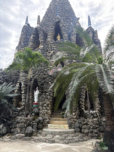 The image captures a unique, ornate structure made predominantly of dark, irregularly shaped stones or coral, resembling a natural grotto or a heavily textured building. Lush green palm trees frame the scene, their fronds extending across the foreground and along the structure, indicating a tropical setting. The sky above is overcast with white and gray clouds.
The structure features multiple spires or pinnacles on its upper levels, some topped with decorative, concentric white discs. Adorning the rock walls are intricate patterns formed by rows of small, white shells, adding a distinct decorative element.
Several figures are visible: a golden statue is nestled within a niche on the upper left side of the structure, while a standing white statue, possibly a religious figure, is positioned closer to the ground on the right. Below the white statue, an entrance or alcove is visible.
To the right of the structure, concrete steps lead up, and a paved path is present. Visible text in green on a concrete surface reads "DE DEP O". Higher up on the rock wall, near the top center, another piece of text, partially obscured and in pink and white, appears to be "CHAMPAL" or a similar word, potentially referencing Cham Pa culture. The scene depicts a static view of this culturally significant and intricately crafted shrine or temple in Cam Ranh, Vietnam.