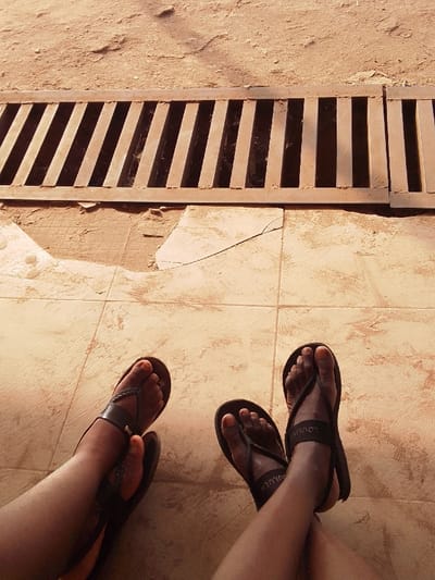 The image displays a ground-level perspective in Nkwelle Ezunaka, Nigeria. Two pairs of dark-skinned human feet, clad in dark flip-flops, are positioned in the lower portion of the frame. One foot features orange-painted toenails, and a sandal strap on the right foot displays the label "LOOLUEN". The feet rest on a light-brown tiled surface, which exhibits cracks and chipped areas. Immediately beyond the tiled section, a horizontal metal grate with diagonal bars spans the width of the image. The ground beyond the grate consists of reddish-brown earth. The scene is illuminated by strong natural light, casting distinct shadows from the grate.