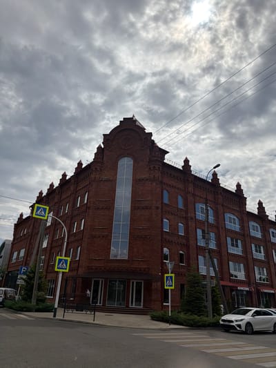The image shows a large, red brick building in Maykop, Russia. The building has many windows, some of which are arched, and decorative architectural elements at the top. In front of the building, there is a street with a crosswalk and a few cars parked. There are also pedestrian crossing signs and a lamppost. The sky is cloudy, with the sun partially visible through the clouds. The overall scene depicts a street view of a historic building in a city.