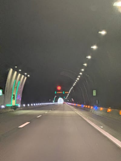 The image shows the interior of a tunnel in Garraf, Spain. The tunnel is lit with rows of lights on both sides. On the left side, the lights are white, while on the right side, they are a mix of orange, blue, and white. Signage is visible on the tunnel walls. There are safety features, including a red circle with "50" indicating a speed limit, and green arrows, suggesting a lane direction or emergency route. There's a road exit indicated on the left. The roadway is gray and appears to be wet or reflective in sections. The image has been rotated counter-clockwise.