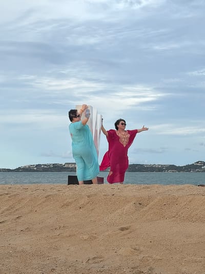 Two individuals are standing on a sandy beach in Ko Samui District, Thailand, with a body of water and a treeline of a hilly island in the background. The sky is overcast with a light gray, cloudy appearance.

In the foreground, on the left, a person wearing a light blue kaftan is taking a photo with their cell phone, holding it up towards a large, white, three-dimensional letter that appears to be the letter "W". The person has dark hair, is wearing sunglasses, and has orange bracelets on their wrist.

To the right of the letter "W", a second person is posing with outstretched arms. This individual is wearing a vibrant pink kaftan with intricate gold embroidery on the chest and sleeves, and is also wearing sunglasses.

The sandy beach has subtle ripples and footprints visible. The water is a calm, blue-green. The island in the distance is covered in dense green foliage and dotted with buildings.
