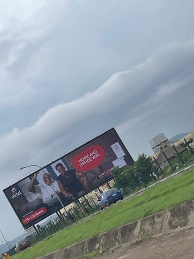 The image shows a billboard advertising Airtel's "Home and Office WiFi" service. The billboard features two men, one in a black shirt and the other in a white shirt, both smiling and appearing to be in a relaxed, positive mood. The text on the billboard is "HOME AND OFFICE WIFI" and includes a phone number "09110000080" and the Airtel logo. The setting appears to be outdoors, with a cloudy sky and some greenery visible in the background, suggesting it might be daytime. In the background, there are also buildings and a road with a blue vehicle. The image is taken from a slightly low angle, looking up at the billboard.
