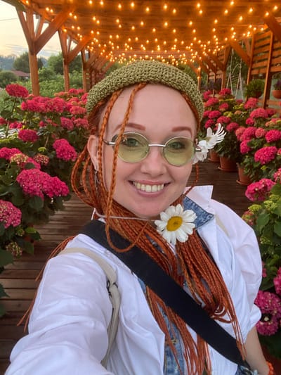 A medium shot portrait shows a woman with red braided hair and a green knitted cap smiling at the camera. She is wearing round sunglasses with green lenses, a white shirt over a denim jacket, and has a daisy and a small white flower in her hair and around her neck. The woman is outdoors, with a wooden pergola overhead adorned with string lights. In the background, there are numerous potted hydrangeas with pink and red blooms. The ground appears to be made of wooden planks. The setting suggests a decorative outdoor seating or dining area.