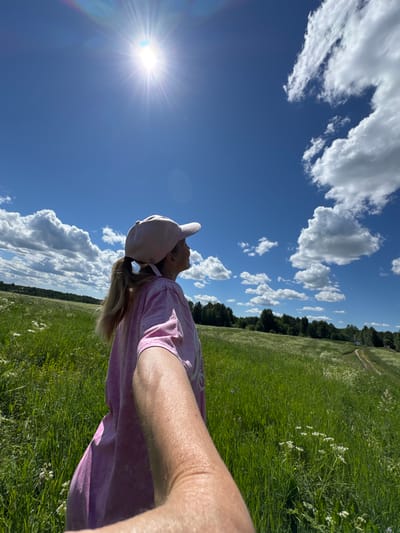 A low-angle shot shows a person wearing a pink t-shirt and a light pink baseball cap, looking up at the bright sun. The person is positioned in the foreground, with their arm and shoulder visible. The sun is radiating light in a starburst pattern, creating a lens flare effect with rainbow colors on the left side of the frame. The scene is a vast, sun-drenched meadow with tall green grass and scattered white wildflowers. In the background, a dense line of dark green trees forms a horizon, with a faint dirt path visible winding through the grass on the right side. The sky is a vibrant blue, dotted with fluffy white cumulus clouds. The overall impression is one of a bright, sunny day in a rural, natural setting, possibly a field or park. The lighting suggests it is daytime, likely late morning or early afternoon given the sun's position and intensity. The person's posture, looking upwards with their head tilted back, could indicate they are enjoying the sun, looking at the sky, or perhaps taking a break. The orientation of the image is vertical, with the subjects and scene appearing sideways.