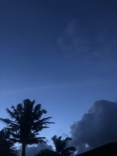 The image captures a twilight scene in Kathaluwa, Sri Lanka, with a deep blue sky dominating the frame. Silhouetted against the darkening sky are several palm trees, their fronds reaching outwards. A single bright star is visible in the upper left quadrant of the sky, just above and to the left of the main palm tree. To the right of the palm trees, large, dark, and ominous storm clouds are gathering, hinting at an approaching weather event. The lower right portion of the image is obscured by the dark silhouette of a rooftop. The overall atmosphere is moody and serene, typical of the transition from day to night in a tropical coastal region.