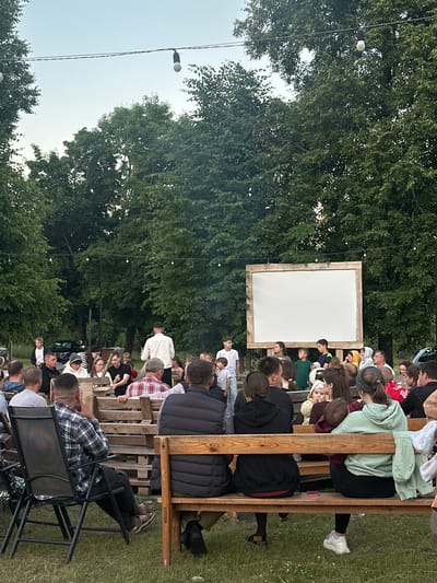 The image depicts an outdoor gathering of people watching a projection screen in what appears to be the evening. The setting is rural, with lush green trees forming a backdrop and a clear sky above. Several strings of lights are visible, suggesting a festive or communal event. A large, blank white screen with a wooden frame is positioned centrally, indicating a movie screening or presentation. A man in a white shirt stands near the screen, possibly addressing the audience. Spectators are seated on wooden benches and chairs, arranged in rows facing the screen. Some individuals are closer to the foreground, while others are further back, creating a sense of depth. The overall atmosphere seems relaxed and convivial. The location is identified as Завалочыцкі сельскі Савет, Belarus.