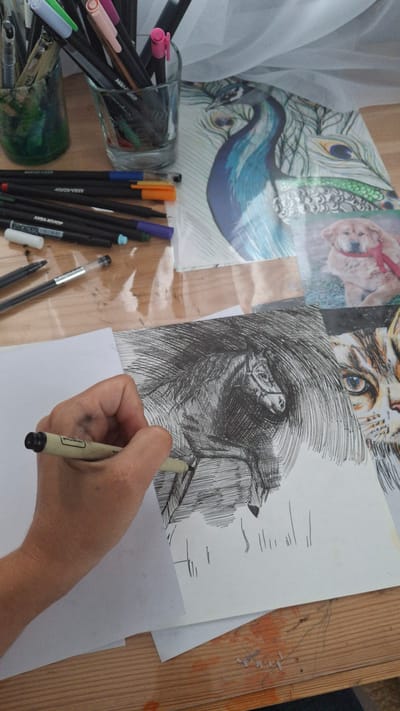 The image shows a person's hand drawing on a piece of paper with a fine-tipped marker. The drawing appears to be a detailed sketch of an animal's eye, rendered with fine lines. Several other art supplies, including markers and pencils in various colors, are scattered on a wooden surface, some in a glass jar. Behind the drawing are collage elements featuring images of peacock feathers and a cat's face. The scene suggests an artistic endeavor, likely taking place indoors given the lighting and arrangement of materials. The overall impression is one of focused creativity. The visible text on the markers is "AKRA-BOJO". No specific time of day, weather, or emotional cues are evident. The context provided indicates the location as Kempelevo, Russia.