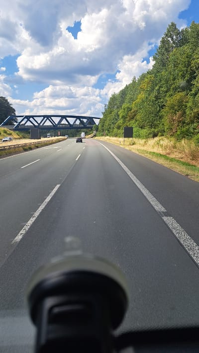 The image shows a view from a vehicle traveling on a multi-lane highway, likely the A45, near Haiger, Germany. A blue truss bridge spans the highway in the distance, with a row of green trees and vegetation lining the right side of the road. The sky is partly cloudy with white cumulus clouds against a blue backdrop. The highway itself is paved asphalt with white lane markings. Several vehicles, including cars and trucks, are visible on the road. In the foreground, a blurry black object, possibly a car mount for a device, is visible.