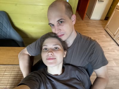 In this close-up, selfie-style photograph, a couple is seated together in what appears to be a domestic setting, possibly a kitchen or dining area, in Ленинградская, Russia. The background features light yellow wood-paneled walls and a light-colored wooden floor. To the left, a dark gray upholstered chair with a diamond-stitched pattern is visible. To the right, a beige refrigerator with a silver handle and a portion of a beige cabinet are present. A wooden door is ajar in the background. The male subject, who is on the left and is bald, is leaning his head towards the female subject. He is wearing a dark gray t-shirt. The female subject, seated in front and embraced by the male, is looking towards the camera with a slight smile. She has light brown hair and is also wearing a dark gray t-shirt. The interaction suggests an intimate and relaxed moment between the couple.