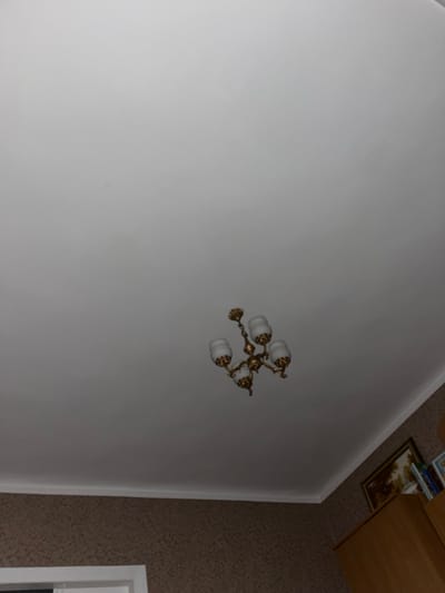 The image shows a ceiling with a vintage chandelier. The chandelier is made of gold-colored metal and has three white glass lamps. The ceiling is white and appears to be made of drywall. Below the ceiling, there is a wall with patterned wallpaper and a white baseboard. In the bottom right corner, a portion of a wooden cabinet and some framed pictures are visible. The scene is indoors, likely in a residential setting.
