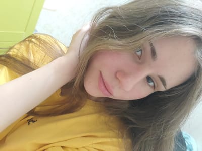 A young woman with long, light brown hair and fair skin is captured in a close-up, selfie-style shot. She is wearing a bright yellow t-shirt with a small, dark embroidered logo on the left side of her chest, which appears to be an abstract symbol or animal. Her right arm is bent, with her hand resting near the back of her head, partially obscuring her hair. Her left arm is extended out of frame. Her head is tilted to her right, and her gaze is directed upwards and slightly to her left, with her eyes clearly visible. Her lips are slightly parted, and she has a subtle, almost imperceptible smile. The background is out of focus, with a pale green surface on the left and a neutral, lighter colored wall on the right. The lighting suggests an indoor environment, possibly a room in Svietlahorsk, Belarus.