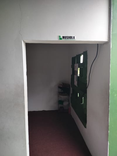 The image displays the entrance to a small, empty room, identified as a "MUSHOLA" by a sign above the doorway. The sign is white with green text and a stylized mosque or minaret icon. The entrance itself is a simple rectangular opening in a white painted wall.
To the left of the entrance, an exterior wall, possibly concrete or stucco, is light gray and shows a distinct vertical crack. On the far right edge of the frame, a vertical section of a green structure or wall is visible, suggesting an exterior element.
Inside the room, the walls are painted white, and the floor is covered with a dark red or brown carpet. On the back wall, a simple, multi-tiered metal shelving unit holds various folded items, which appear to be fabrics or clothes, along with some round objects resembling plates or bowls. A small red object, possibly a fire extinguisher, stands on the floor near the shelf.
The right interior wall features a section painted dark green, composed of horizontal slats or panels with small rectangular openings, through which natural light appears to enter. A black electrical cable runs vertically down this green section. The overall lighting suggests it is daytime. The room is unoccupied and appears to be a functional, modest prayer space, consistent with the "MUSHOLA" designation in the Dramaga, Indonesia context.