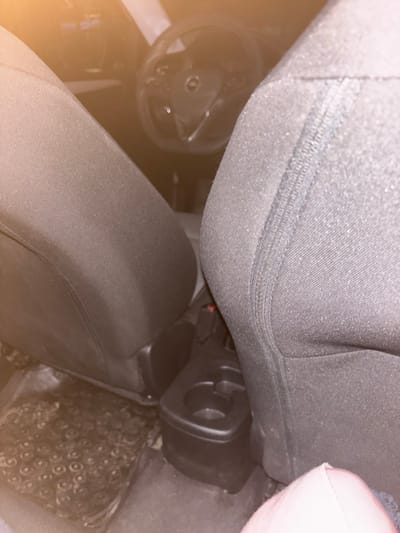 The image depicts the interior of a stationary vehicle in Novo Panicharevo, Bulgaria, viewed from a rearward position looking towards the front. The backs of two dark-colored, fabric-upholstered front seats occupy the mid-ground, framing a central gap. Between the seats, a dark-colored plastic center console is visible, featuring at least one circular cup holder. A red seatbelt release button on a seatbelt receptacle is discernible near the passenger-side seat base. On the driver's side floor, a dark, textured rubber floor mat is present. In the background, beyond the front seats, the vehicle's steering wheel is partially visible, featuring a central emblem and control buttons on its spokes. A portion of a light-colored, possibly pink fabric object, is present in the lower right foreground. The scene is dimly lit, with a prominent warm, yellowish lens flare originating from the upper left, contributing to an overall low-light environment. No individuals are visible.