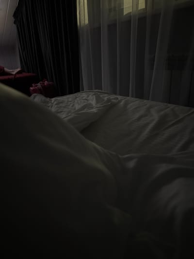 The image depicts a dimly lit bedroom. A bed with white sheets is prominent in the foreground, appearing rumpled. In the background, dark curtains hang on the left, and sheer white curtains are visible on the right, allowing some light to filter through from outside. A radiator can be seen behind the sheer curtains. On a piece of furniture to the left of the bed, there is a red pillow or cushion with a person's legs resting on it, and a pink bag or piece of clothing is also present. The overall lighting suggests it may be early morning or late evening. The location is Doma 1096 Km, Russia.