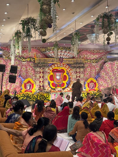 The image depicts an indoor gathering within a large hall in Bidhannagar, India, focused on an elaborately decorated stage or altar. The high ceiling features recessed lighting, suspended decorative wicker spheres, and garlands of white artificial flowers and green foliage. A large crystal chandelier is partially visible on the right.

The central feature of the scene is a wide stage adorned with a vibrant, intricate backdrop displaying pink, gold, red, and yellow patterns. At the center of the stage is a prominent, ornate shrine or religious effigy, heavily decorated with red, yellow, and blue flowers, encased in a gilded frame. Flanking this central shrine are two smaller, lotus-shaped floral arrangements, each containing an image or deity. A platform covered with yellow flowers extends across the stage foreground. On the left side of the stage, two black professional audio speakers are visible.

Numerous individuals are present, predominantly seated on the floor on white sheets facing the stage. A section of orange upholstered couches is visible on the far left, also occupied by seated individuals. The attendees are primarily adults, many wearing traditional Indian attire such as saris in various colors including red, orange, pink, and yellow. Several individuals in the foreground are observed holding open books or religious texts. On the stage, several men are present. One man in a brown uniform stands with his back to the viewer near the central shrine, while other men are seated or standing on the stage, some in traditional attire. The overall scene suggests a religious or cultural ceremony or event.