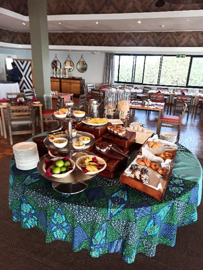A buffet table displays assorted breakfast items. Doughnuts, pastries, and cereals are arranged in containers. A tiered stand holds small bowls of yogurt and fruit. Plates are stacked at the edge of the table. A colorful, patterned tablecloth covers the buffet table. Chairs and tables extend into the background, alongside a large window with a view of greenery.