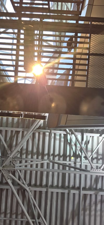 The image shows a view looking up towards the ceiling of a large, open space, likely an airport terminal, in Vnukovo, Russia. The ceiling is composed of multiple levels of industrial-style architecture. The upper level features a translucent, possibly glass, roof with a grid-like structure of dark beams. Sunlight streams through this roof, creating bright glare and lens flare from a spotlight. Below this, there is a darker, horizontal beam with reflective surfaces, upon which a black, silhouetted figure, possibly a person, is visible in the upper part of the frame. Further down, the ceiling transitions to corrugated metal panels, supported by a complex network of silver metal trusses and beams. The overall impression is one of an expansive, modern, and possibly recently constructed indoor space.