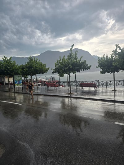The image depicts a rainy day in Lugano, Switzerland, with a focus on a waterfront promenade. The scene is dominated by a dramatic sky filled with dark, heavy clouds, hinting at recent or ongoing rainfall. In the background, imposing mountains rise, their peaks partially obscured by the overcast conditions.
The promenade itself is wet and reflective, showcasing the effect of the rain. Several red benches are visible along the waterfront, sparsely occupied, suggesting a quiet, perhaps somber, atmosphere due to the weather. Trees with lush green foliage line the promenade, their leaves glistening with moisture. A metal railing with an ornate design separates the walkway from the body of water, which appears calm and grey under the moody sky.
In the foreground, two individuals are captured mid-stride on the wet pavement. They appear to be running or walking briskly, possibly seeking shelter or enjoying the unusual conditions. One person, wearing a dark tank top and shorts, is more prominently visible, her hair wet from the rain. The other person, in lighter clothing, is partially obscured. The overall mood is one of a tranquil yet slightly melancholic day, with nature's elements taking center stage.
A sign with text is partially visible near the people, featuring a colorful image and French text, possibly an advertisement or informational display. The time of day is indeterminate due to the cloud cover, but the low light suggests it could be late afternoon or early morning. The reflections on the wet asphalt add a sense of depth and visual interest to the composition, highlighting the pervasive moisture.