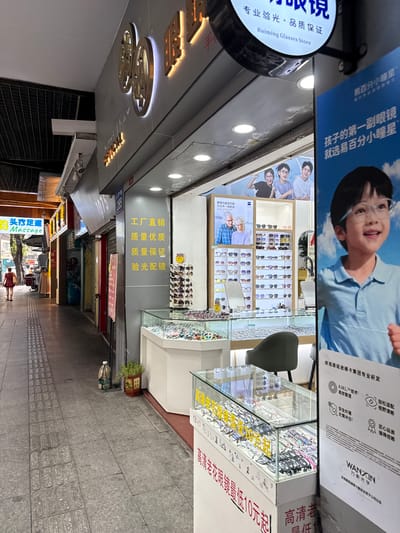The image shows a street-level view of an optical shop in Yuexiu District, China. The shop's facade is illuminated by bright signage, with large golden characters and a circular logo that reads "Ruiming Glasses Store" in English. Inside, the shop displays a variety of eyeglasses and sunglasses on shelves and in glass cases. A large poster features a young boy wearing glasses, and other advertisements are visible. The street outside is paved with grey tiles, and a woman in a red dress is walking away from the camera. The overall scene depicts a typical commercial street in an urban Chinese setting, with a focus on retail and consumer goods.