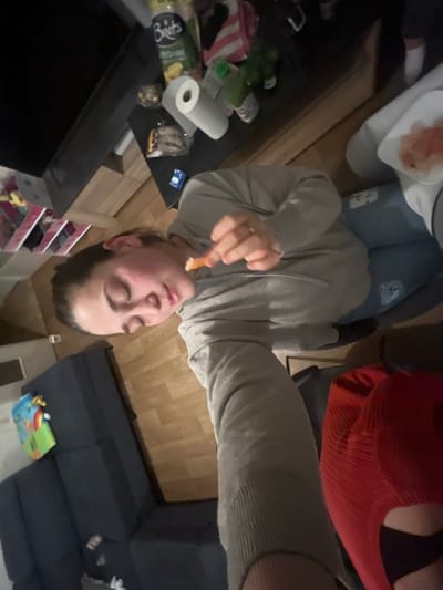 A person in a grey hoodie holds an orange object to their mouth. The scene is indoors. To the left, a dark surface holds paper towels and food containers. Further in the background, a shelf unit with children's items. A blue sectional couch is in the lower right corner, with a colorful mat in front of it. The photo is taken at an angle, appearing tilted.