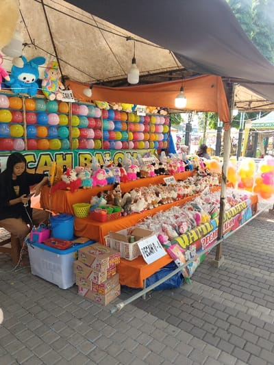 This image captures an outdoor game stall, likely at a market or fairground, in Ciamis, Indonesia. The main subject is a "Pop the Balloon" game booth, identified by the prominent "PECAH BALON" text (Indonesian for "Pop the Balloon") and rows of colorful balloons arranged in a grid for players to aim at.

One young woman, presumably the vendor, is seated on a stool to the left, looking down at her phone. She is wearing a dark top and light-colored pants. Next to her are a blue cooler, a pink container, and a basket.

The stall is covered by a dark and orange awning, with several exposed lightbulbs hanging for illumination. Below the balloon wall, a long table displays numerous prizes. These include a vast array of plush toys and stuffed animals, such as bunnies, cartoon characters, and various other animals, organized on tiered orange fabric. Many smaller prizes, like packaged toys or souvenirs, are also visible in plastic bags, hanging or arranged on the lower sections of the table.

White signs with "PECAH" (meaning "Pop" or "Broken") followed by alphanumeric codes like "PECAH B-1" and "PECAH A-4" are placed amongst the prizes, likely indicating prize tiers or winning conditions. In front of the table, stacked cardboard boxes of "Teh Gelas" (an instant tea brand), marked "100% ASLI" and "PACK", are visible on the ground.

The scene takes place during bright daylight, suggested by the clear shadows and overall illumination, though the string lights under the awning indicate potential evening operation. The ground is paved with interlocking concrete blocks. In the background, beyond the stall, trees and other market structures are vaguely visible, including a partially legible sign that reads "BUBUR INSTAN" (instant porridge). The atmosphere appears casual and lively, typical of a local outdoor event.