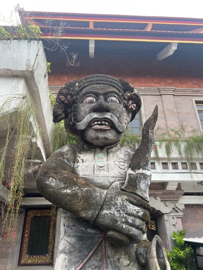 A weathered stone statue of a male figure with an exaggerated expression, featuring bulging eyes, a large, curled mustache, and a wide grin, stands in Celuk, Indonesia. The statue wears a stylized armor or garment with decorative elements. The figure appears to be raising its right arm in a gesture. The backdrop shows a multi-story building with mixed architectural styles, including brick and modern design. Vegetation, including small-leaved plants and red flowers, is visible around the base of the statue. The image is oriented vertically, with the statue dominating the foreground.