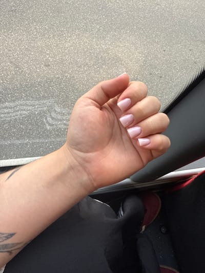 An overhead, close-up shot shows a person's left hand with light pink, square-shaped fingernails. The hand is positioned in the foreground, with the palm facing upwards. Behind the hand, a textured gray surface, possibly pavement, is visible. A black dashboard or car interior occupies the right side of the frame, with a sliver of a car's windshield visible along the top edge. The person's arm extends into the lower left portion of the image, showing a faint tattoo on the forearm. The background suggests an outdoor setting, likely within the city of Breisach am Rhein, Germany. The lighting is natural, indicating daytime.