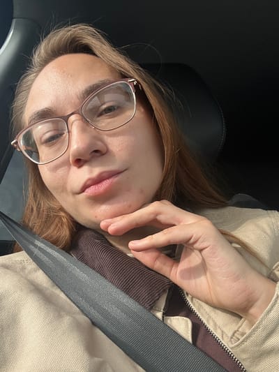 A young woman with light brown hair and fair skin is in a car, wearing glasses and a seatbelt. She has on a tan jacket and a brown corduroy shirt. The car's interior is dark, and the image appears to be taken inside the vehicle. The location in the city of Kvarsa, Russia cannot be determined from the image.