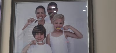 The image displays a silver-framed picture hanging on a light beige wall in Kogon City, Uzbekistan. The photograph within the frame depicts a smiling family of four: an adult woman, an adult man, a young girl, and a young boy. All four individuals are holding toothbrushes near their mouths, simulating the action of brushing. The adult woman and man are positioned in the background, both wearing white attire resembling bathrobes or medical coats. The woman holds a light blue/green toothbrush in her right hand. The man, positioned slightly behind her, holds a yellow toothbrush in his left hand. In the foreground, the young girl wears a white tank top and holds a blue and yellow toothbrush in her right hand. The young boy, positioned to the left of the girl, wears a white t-shirt and holds a light blue/green toothbrush in his right hand. The background of the photo itself is a plain light grey or white. Significant reflections are present on the glass surface of the framed picture, including horizontal lines consistent with window blinds and several bright white light sources, particularly prominent on the man's forehead and the upper right corner of the frame. A faint purple reflection is visible along the left vertical edge of the framed picture.