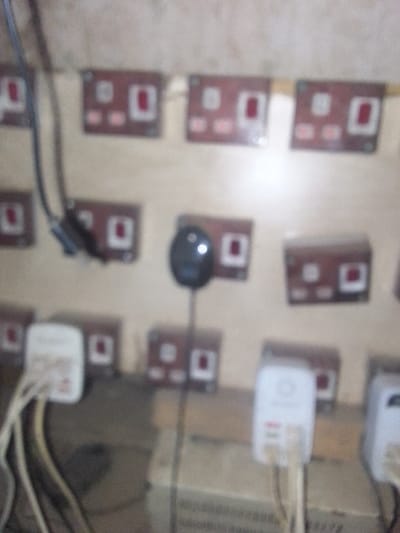 A collection of brown British wall sockets are arranged on a light brown wall, some occupied by white plugs with visible white cables. A black, bulb-shaped object is plugged into one of the sockets. The scene appears to be poorly lit and slightly out of focus.