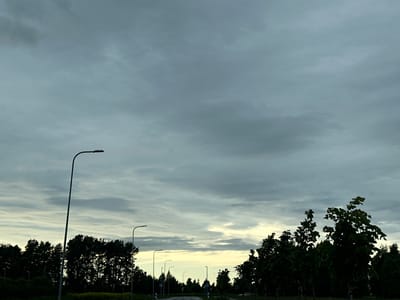 The image depicts a road in Tiskre, Estonia, under a cloudy, overcast sky. The scene is dominated by a dramatic sky with heavy, textured clouds that create a somber and moody atmosphere. The time of day appears to be late afternoon or early evening, given the soft, diffused light.

Along the left side of the road, a tall, modern streetlamp stands prominently, its curved arm extending over the roadway. Further down the road, a series of shorter streetlamps are visible, spaced at intervals, suggesting this is a well-lit path or road.

On either side of the road, dense foliage and trees form dark silhouettes against the lighter sky. The trees on the left appear to be a thick forest or line of trees, while those on the right are more spread out, with some taller trees featuring broad leaves catching the muted light. The ground is covered in greenery, with some darker, shadowy areas indicating the presence of bushes or dense undergrowth.

The road itself curves gently into the distance, disappearing into the horizon. There are no people visible in the image. The overall impression is one of quietude and perhaps a hint of melancholy due to the weather and the subdued lighting. The natural elements of trees and sky are juxtaposed with the man-made elements of the road and streetlamps. No visible text is present in the image.