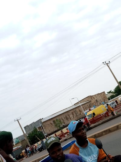 This image captures a street scene in Tumfure, Nigeria, likely during the day under an overcast sky. The foreground features three individuals, two of whom are prominently visible. One man, wearing an orange and light blue shirt and a blue baseball cap, is looking to the right. The cap has a logo with the words "MAYOR" and an emblem. Another man, wearing a green beret, is partially visible on the left, looking to the right. A third person, in a purple shirt, is also in the foreground, though less distinct.
In the background, several people are gathered, some near motorcycles and a dark-colored car. There's a yellow van with writing on its side, and a building with air conditioning units and satellite dishes. A prominent utility pole with multiple wires stretches across the upper portion of the frame, indicating an urban or semi-urban setting. Red fencing lines a pathway or road. One of the men in the foreground is wearing a green cap with white text that appears to read "AHMAD YUGUDA 27". The overall atmosphere seems to be one of everyday life or perhaps a gathering related to a local event or political campaign.