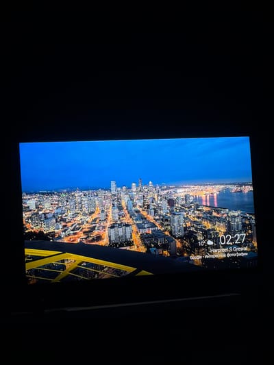 The image displays a nighttime cityscape viewed from a high vantage point, possibly from a tall building or a bridge. The sky is a deep blue, transitioning to black at the top. The city below is illuminated with a myriad of lights from buildings and streetlamps, creating a vibrant and dense urban landscape. A body of water, likely a bay or river, is visible on the right side of the image, reflecting the city lights.

In the foreground, a prominent yellow structure, resembling a bridge or a railing, spans across the lower part of the image. The screen shows a time display of "02:27" and weather information indicating "10°". Below that, text reads "Harpreet S Grewal" followed by "Google • Интересные фотографии". This suggests the image is a screenshot or a photo taken from a device displaying a city view, possibly a live wallpaper or a photo application.

The provided information suggests the image is related to the city of Lysva, Russia. However, the visual elements in the image - the dense, modern cityscape with a waterfront and specific architectural styles - do not immediately correspond to typical imagery of Lysva, which is a smaller industrial city. Without additional context or visual cues that specifically identify Lysva, it is not possible to definitively confirm its presence in the image based solely on the visual data. The image strongly resembles a view of a larger, more developed coastal city.